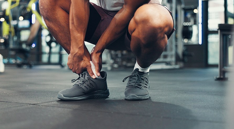 En man sitter p� huk i gymmilj� och h�ller h�nderna runt sin fotled.